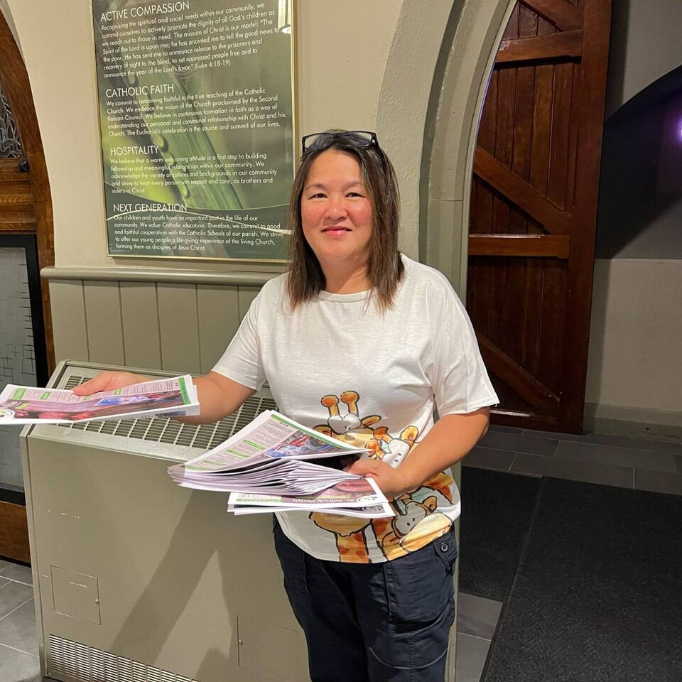 A smiling parish volunteer at the church entrance hands out the weekly bulletin, beneath a plaque of the parish values